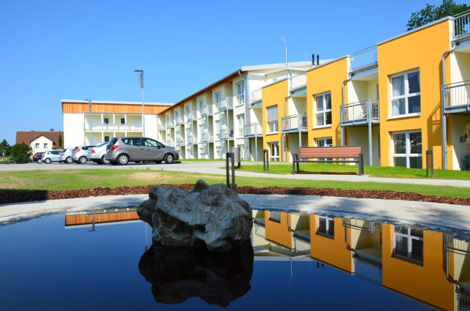Modernes Wohngebäude mit Wasserteich und Garten, sonniger Tag, Parkplätze vorhanden.
